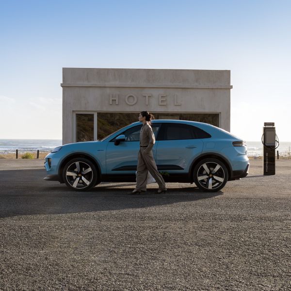 Side shot of a Frozen Blue Metallic Macan Electric parking in front of a modern building by the beach with a woman standing in front of the car.