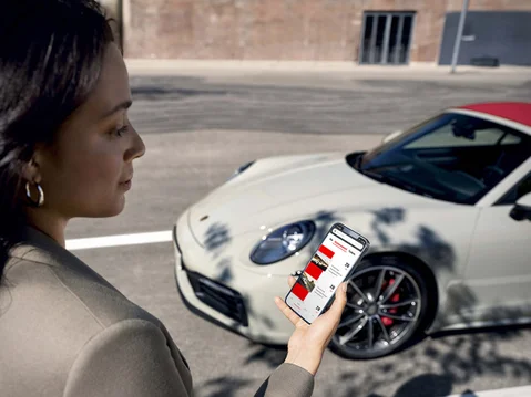 Een vrouw staat voor een Porsche met een mobiel in haar hand, hierop is de Porsche 24 app te zien.