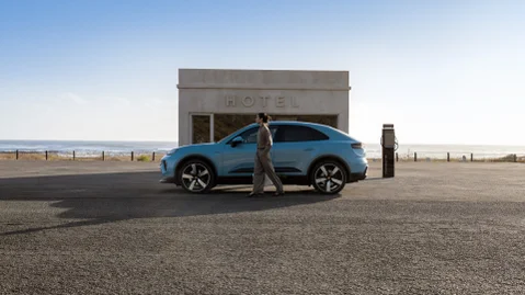 Side shot of a Frozen Blue Metallic Macan Electric parking in front of a modern building by the beach with a woman standing in front of the car.
