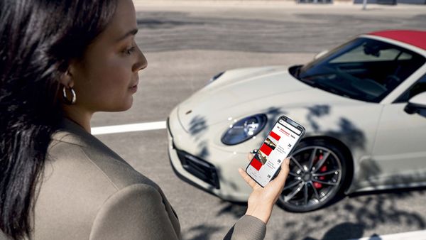 Een vrouw staat voor een Porsche met een mobiel in haar hand, hierop is de Porsche 24 app te zien.