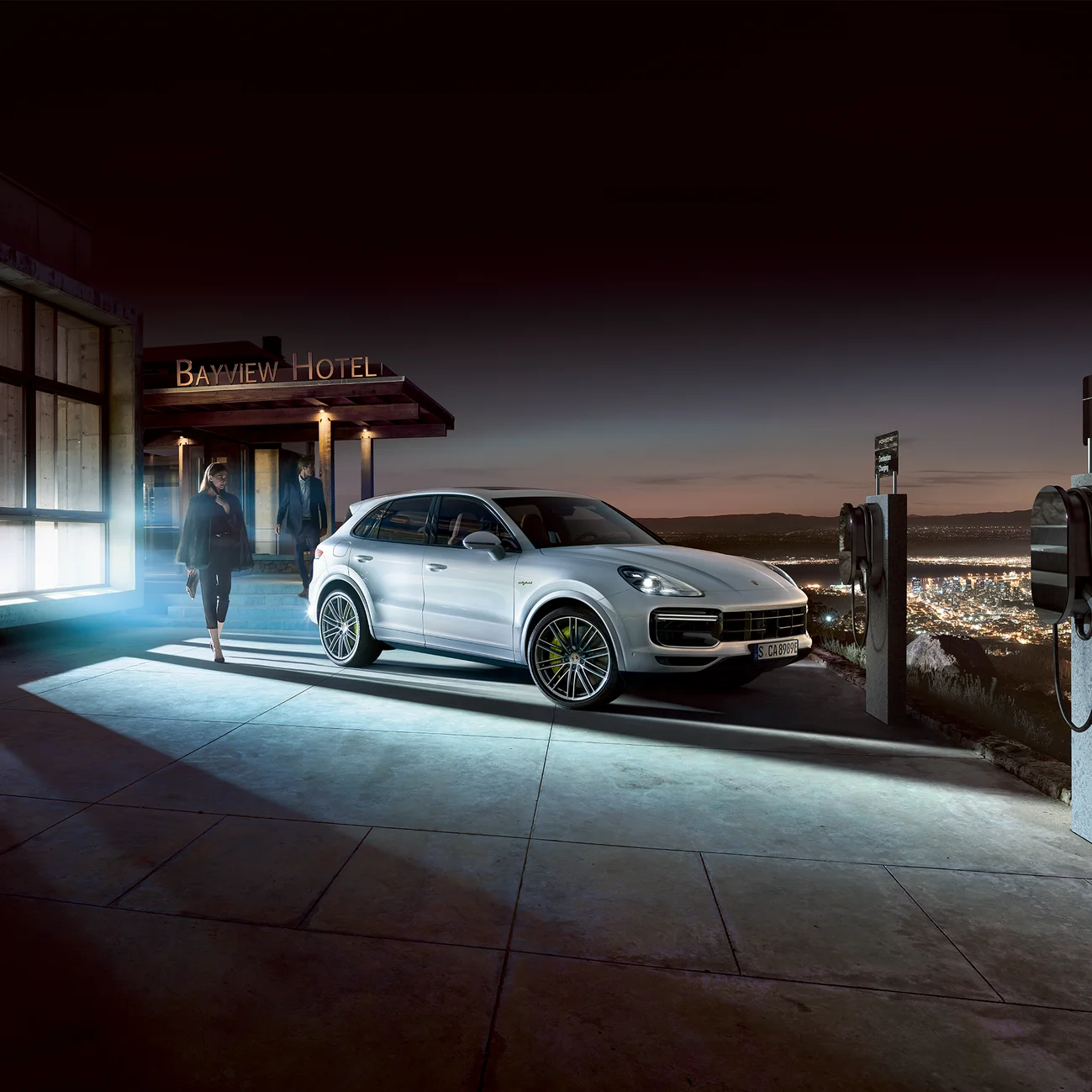A woman waiting for Cayenne E-Hybrid to be charged next to a electric car charger in front of the hotel.