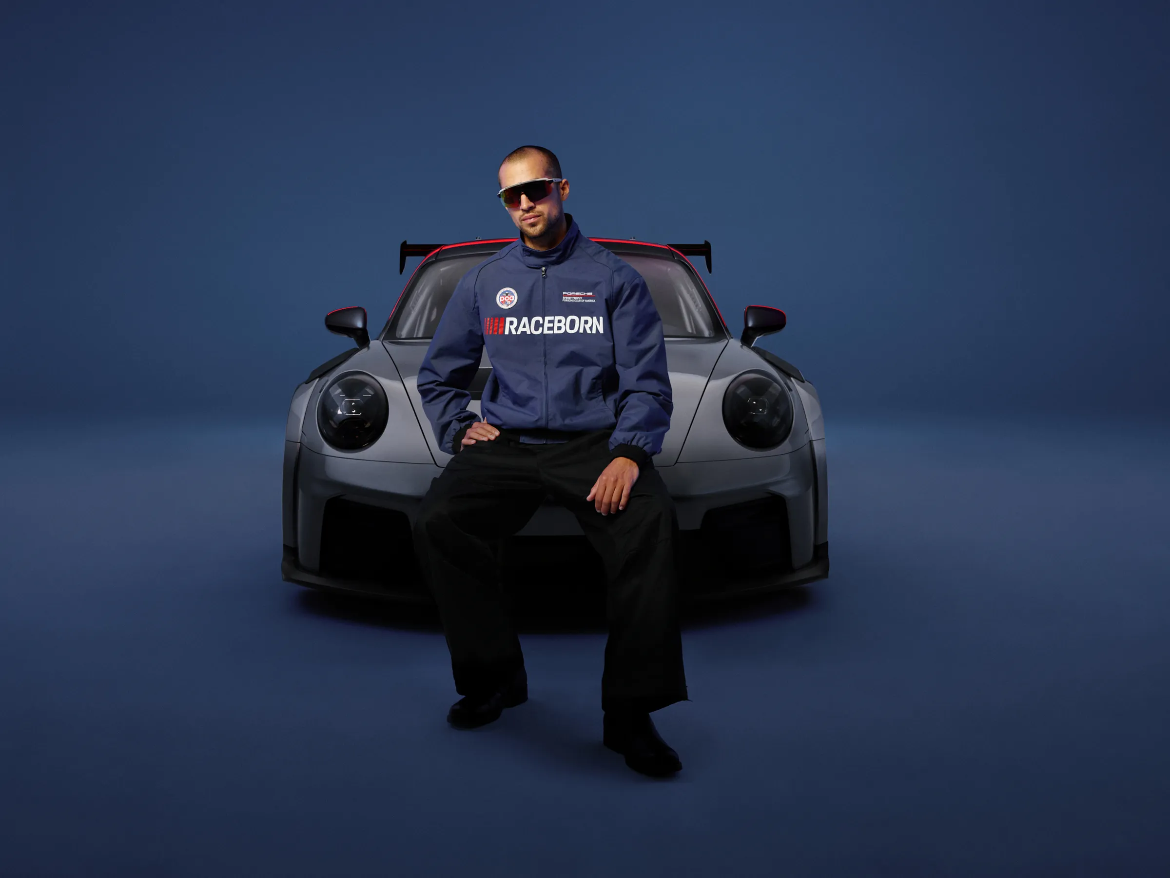 Person wearing sunglasses, black pants, and a blue Porsche "RACEBORN" jacket sits on the front of a gray Porsche 911 GT3 RS, against a dark blue studio background.