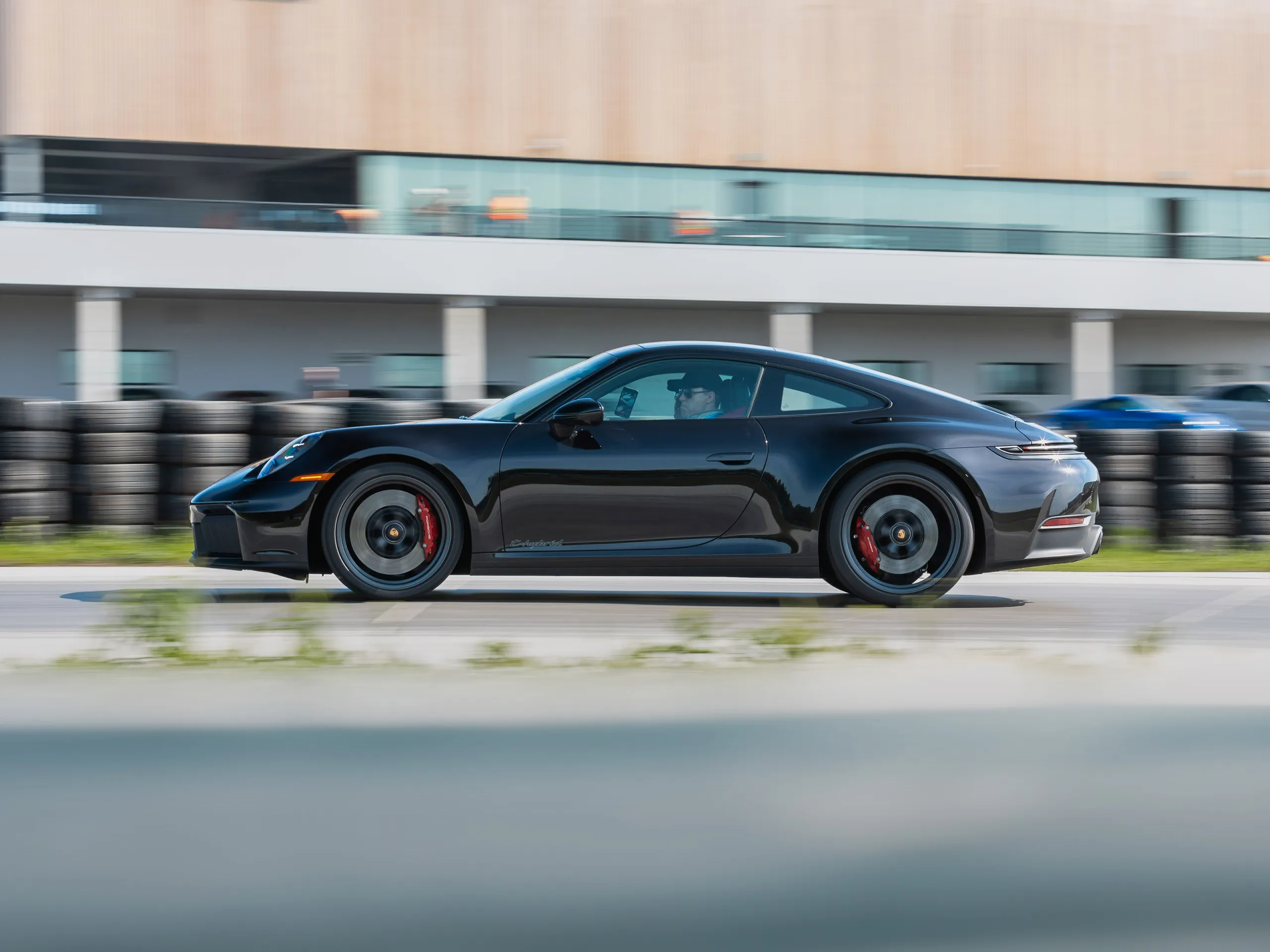A moving shot of a black Porsche 911 driving down a track with a modern building in the background.