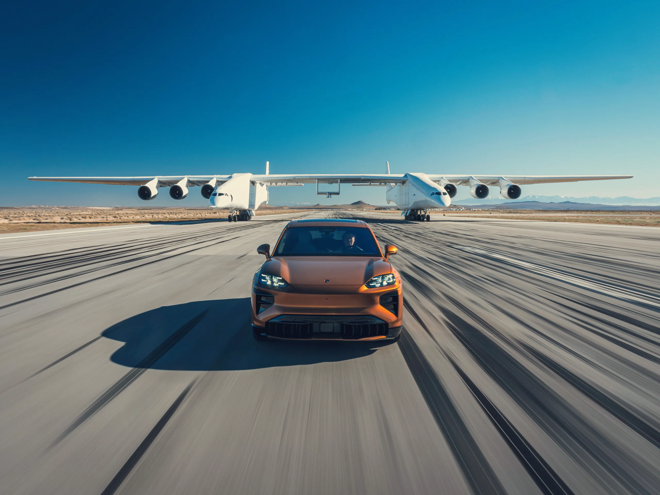Porsche Cayenne Electric vehicle driving on an airfield with a Stratolaunch Roc airplane following behind.