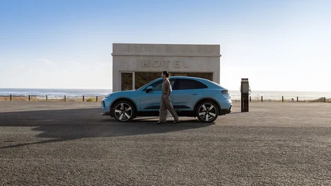 Side shot of a Frozen Blue Metallic Macan Electric parking in front of a modern building by the beach with a woman standing in front of the car.